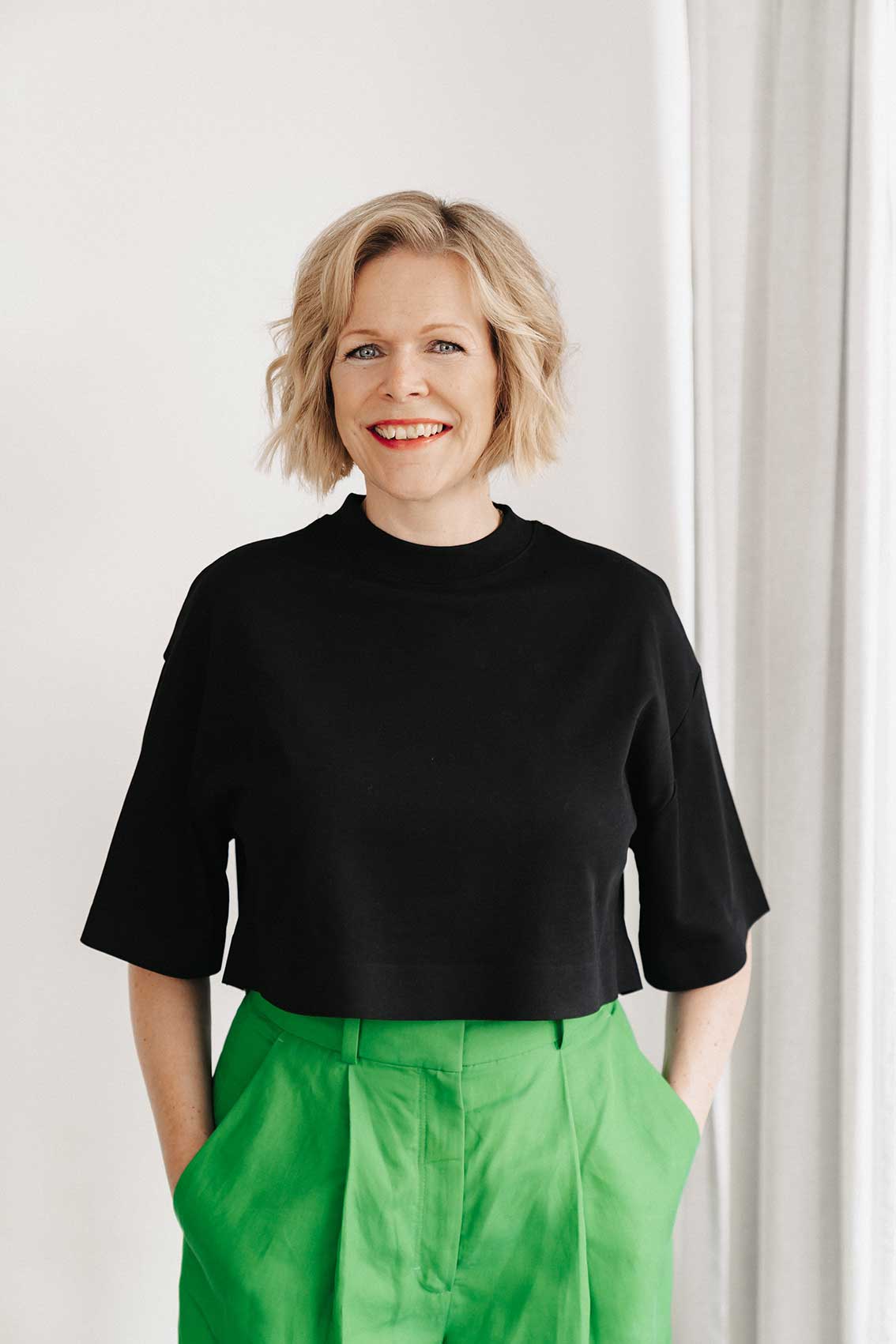 Portrait von Britta im schwarzen Shirt und grüner Hose, stehend, lachend, Blick in die Kamera