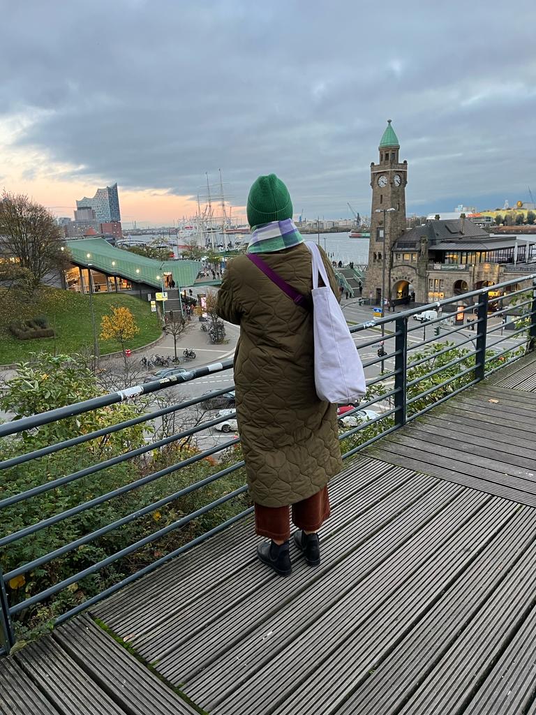 Britta steht auf einem Aussichtspunkt und schaut auf die Landungsbrücken Hamburg. Es ist Herbst und der Tag neigt sich dem Ende zu.